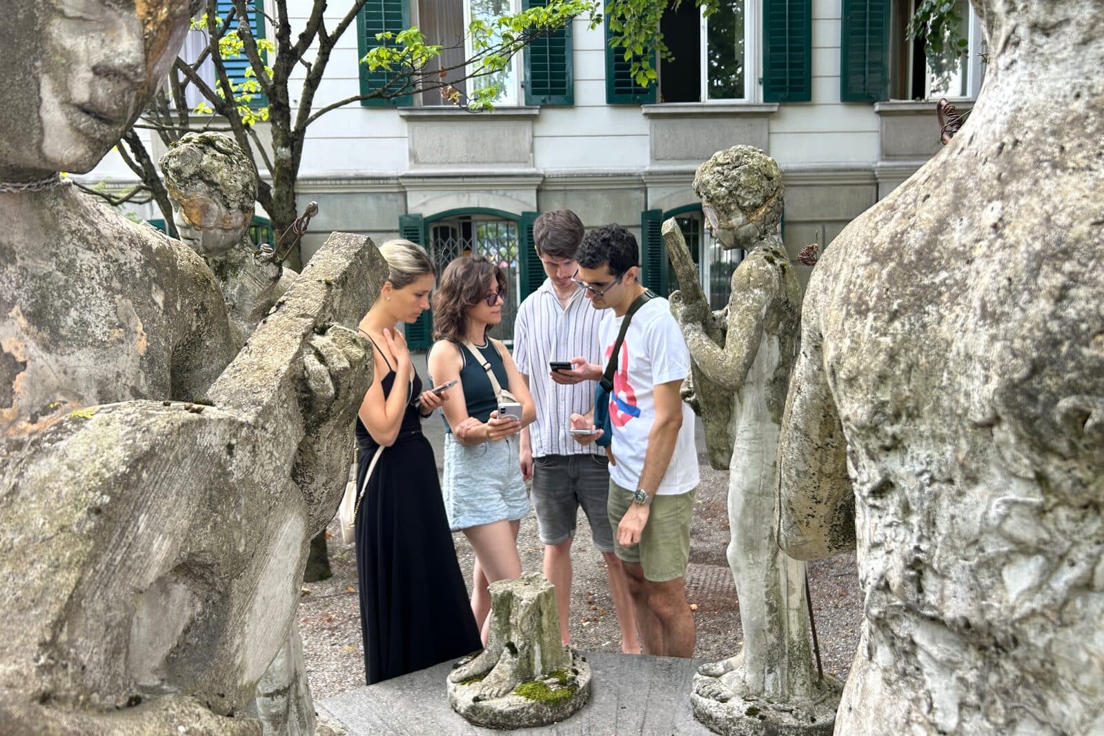 Four players standing among the stone figures of the Eight Musicians sculpture in Zürich, checking clues on their phones.