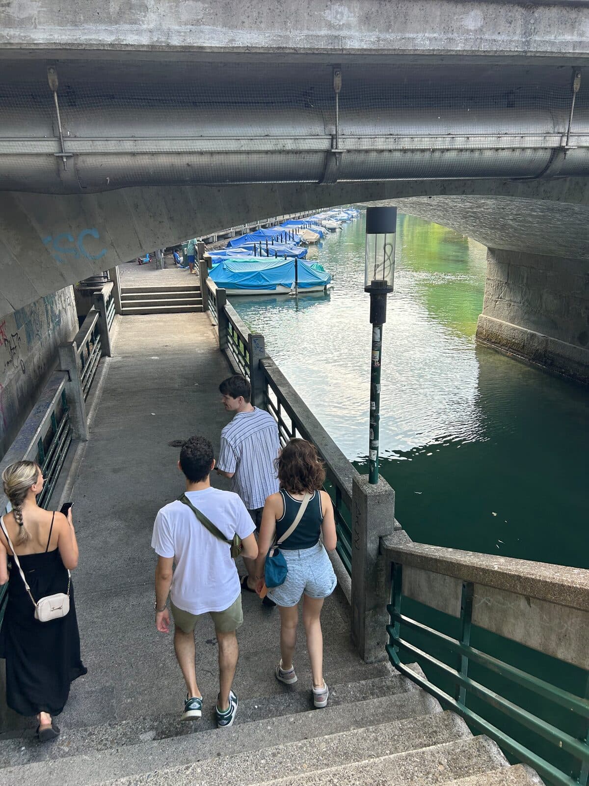 A group walking down steps under a bridge along the Schanzengraben canal in Zürich, moored boats ahead of them.