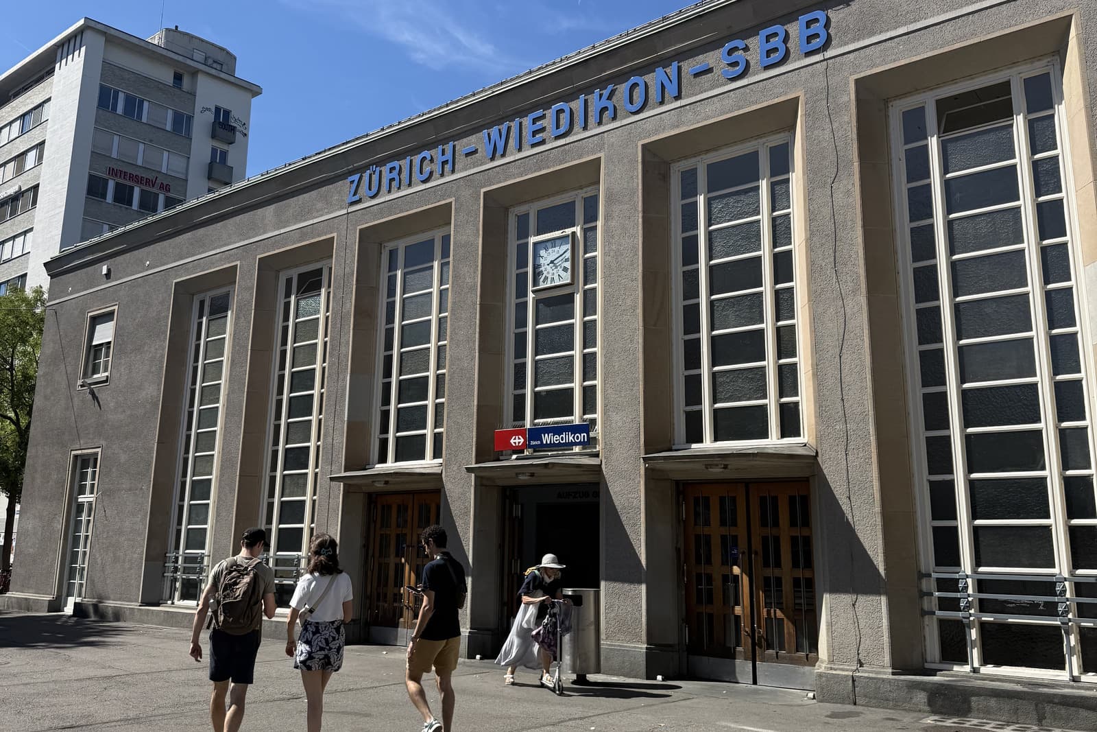 Three players approaching the grey Zürich-Wiedikon SBB station, blue station name across the facade.